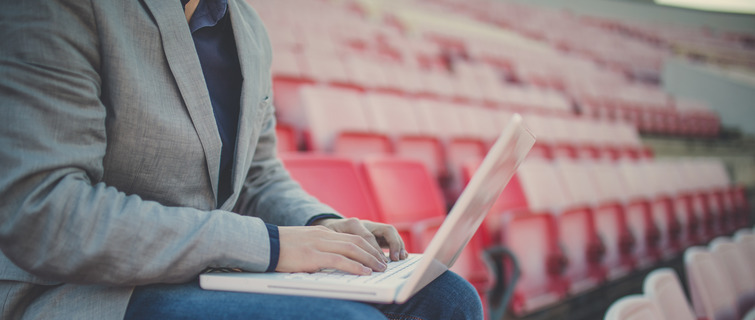 Young bearded coach sitting on the stadium looking at his laptop.