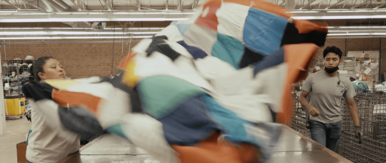 Two workers unfolding a large, colorful quilt-like textile in a workshop